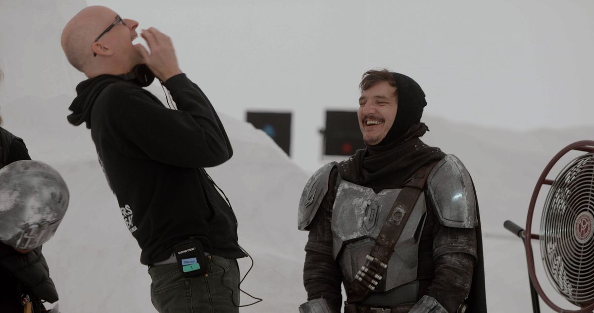 Peyton Reed et Pedro Pascal rient sur le tournage de la saison 2 de The Mandalorian. Pascal porte son costume, mais sans casque, une cagoule noire lui couvrant la tête et décoiffant ses cheveux.