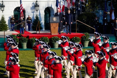Des soldats vêtus de manteaux rouges et d'uniformes militaires du XVIIIe siècle défilent devant une scène où se tiennent Joe Biden et Emmanuel Macron.